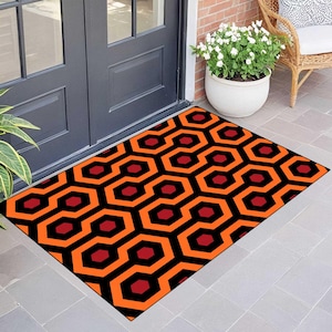 Op de afbeelding: Rechthoekige deurmat met een herhalend geometrisch patroon in zwart, oranje en rood. Het ontwerp bevat onderling verbonden zeshoeken en achthoeken. De mat ligt voor een donkergrijze deur op een betegeld oppervlak.