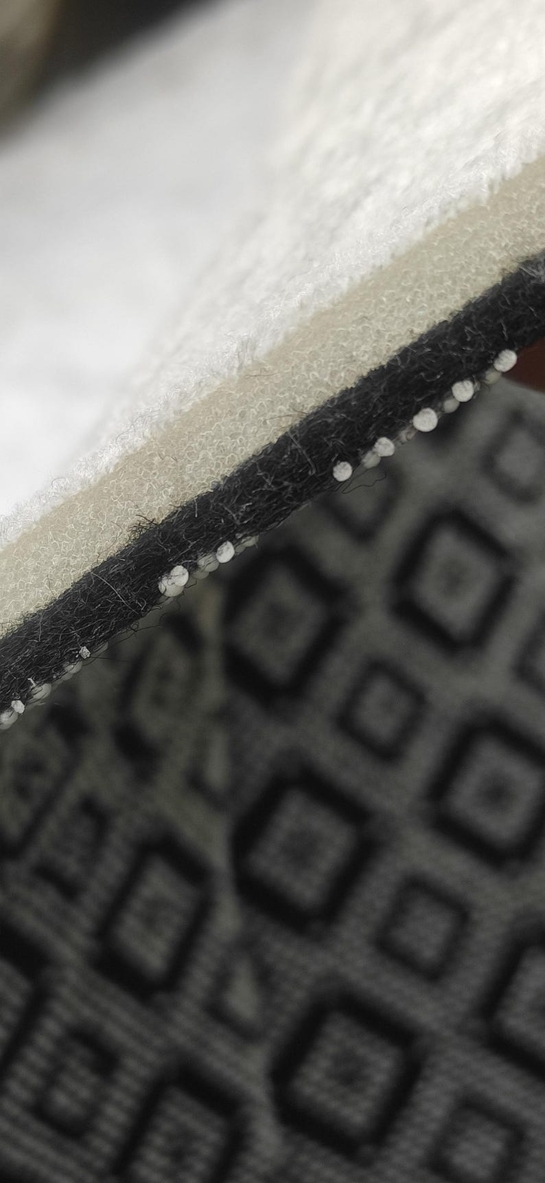 May include: Close-up of a layered textile with a geometric pattern. The top layer is white, followed by a beige layer, and a black layer with white stitching. The bottom layer features a repeating diamond pattern in shades of gray.