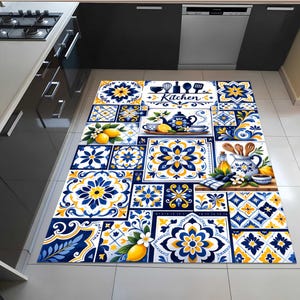 Op de afbeelding: Een rechthoekig keukenkleed met een blauw, geel en wit tegelpatroon. Het kleed heeft bloemen- en geometrische ontwerpen, citroenen, kopjes en het woord "Kitchen". Het kleed ligt op een betegelde vloer.