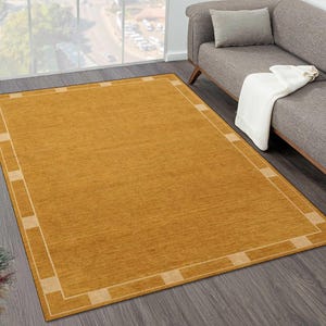 May include: A rectangular area rug in a solid mustard yellow color featuring a lighter beige border with a repeating block pattern. The rug has a flat, woven texture and is positioned on a dark wood floor in front of a gray sofa with a white throw.