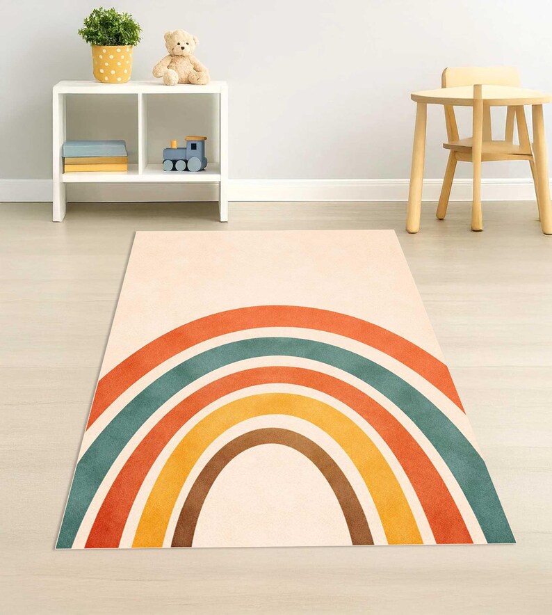 May include: A rectangular area rug featuring a centered, multi-colored rainbow graphic. The rainbow consists of concentric semi-circular bands in shades of burnt orange, muted teal, golden yellow, and dark brown against a cream-colored background.