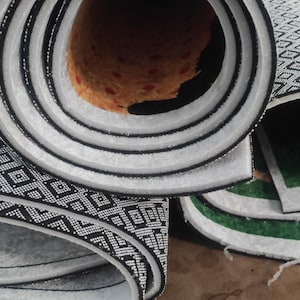 May include: A rolled-up rug featuring a black and white geometric pattern on the underside. The interior of the roll reveals a textured orange backing with small red circular accents, while the edges show a thick white foam layer.