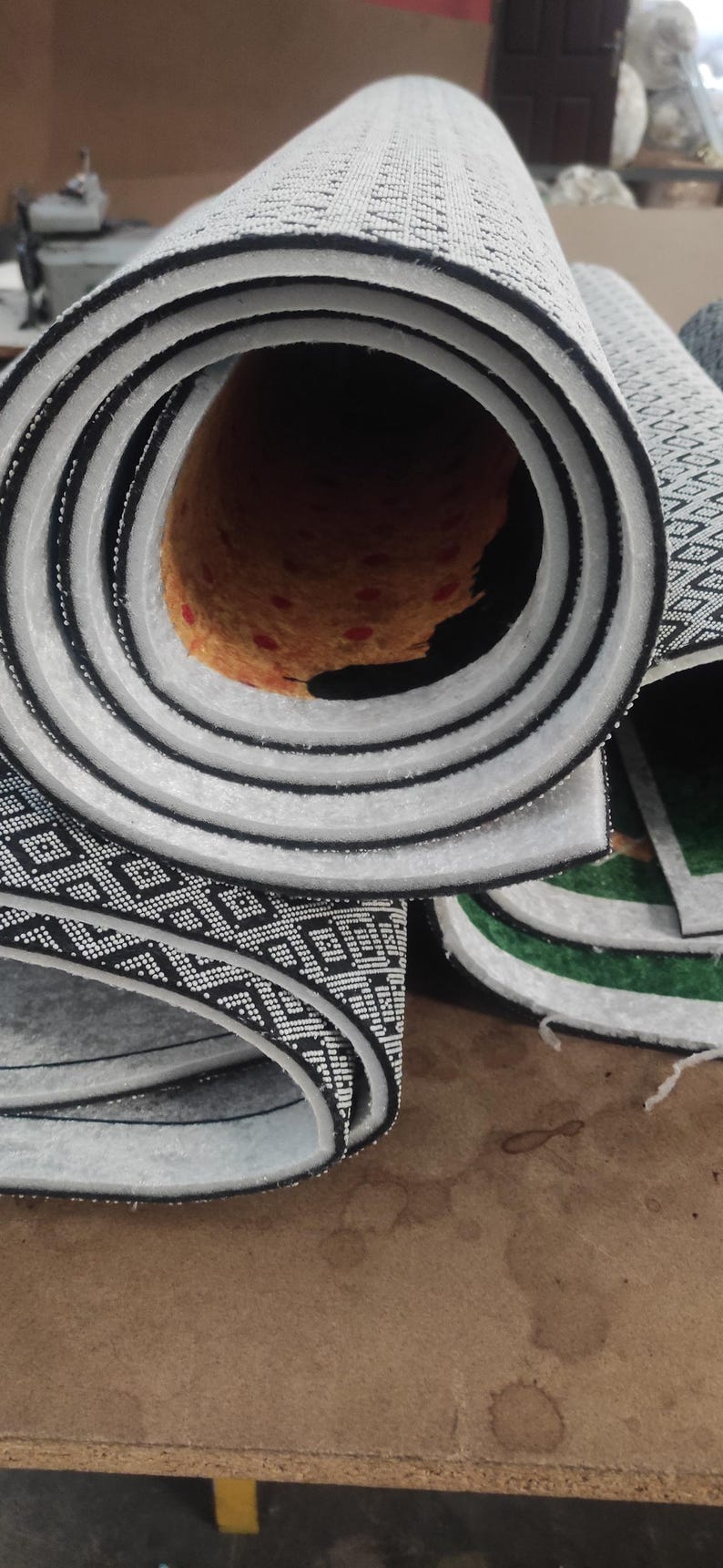 May include: A rolled-up rug featuring a black and white geometric pattern on the underside. The interior of the roll reveals a textured orange backing with small red circular accents, while the edges show a thick white foam layer.