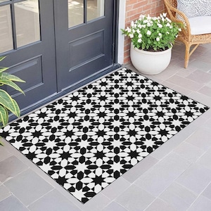 May include: Black and white geometric patterned doormat. The mat features a repeating starburst design. The doormat is rectangular and placed in front of a dark gray door. A white flower pot and wicker chair are in the background.