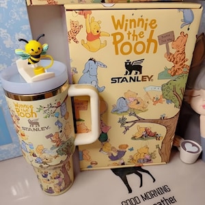 May include: A Winnie the Pooh themed Stanley travel mug with a yellow and blue bee-shaped lid ornament. The mug and box feature illustrations of Winnie the Pooh and friends. The box and mug have the text "Winnie the Pooh" and "Stanley".