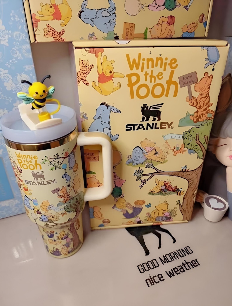 May include: A Winnie the Pooh themed Stanley travel mug with a yellow and blue bee-shaped lid ornament. The mug and box feature illustrations of Winnie the Pooh and friends. The box and mug have the text "Winnie the Pooh" and "Stanley".