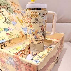 Könnte beinhalten: Ein beigefarbener Stanley-Becher mit Winnie-the-Pooh-Figuren, einem weißen Griff und Deckel sowie einem gelben Bienen-Topper. Der Becher befindet sich in einer passenden Box mit Illustrationen derselben Charaktere.