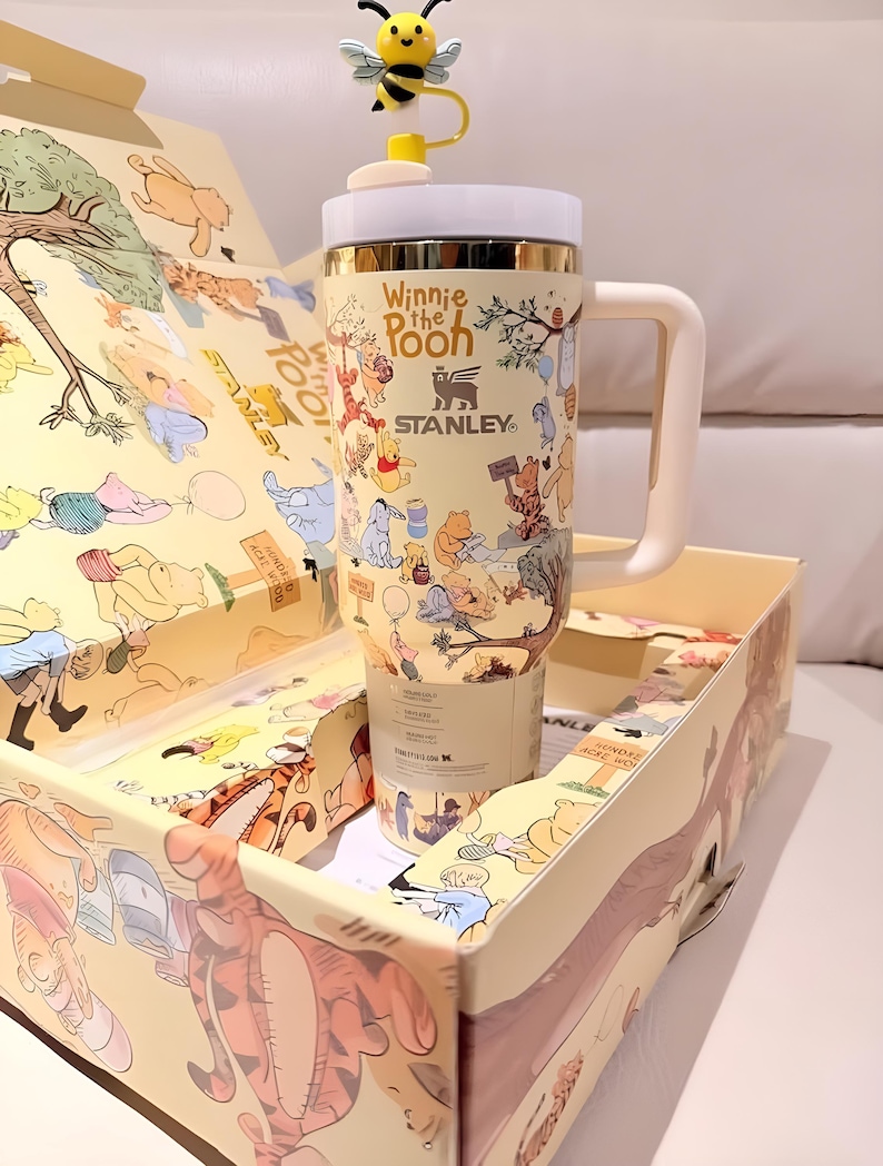 May include: A beige Stanley tumbler featuring Winnie the Pooh characters, with a white handle and lid, and a yellow bee-shaped topper. The tumbler is inside a matching box with illustrations of the same characters.