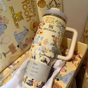 May include: A yellow Stanley tumbler featuring Winnie the Pooh characters. The tumbler has a white handle and a light blue lid. The tumbler is in a yellow box with matching Winnie the Pooh illustrations.