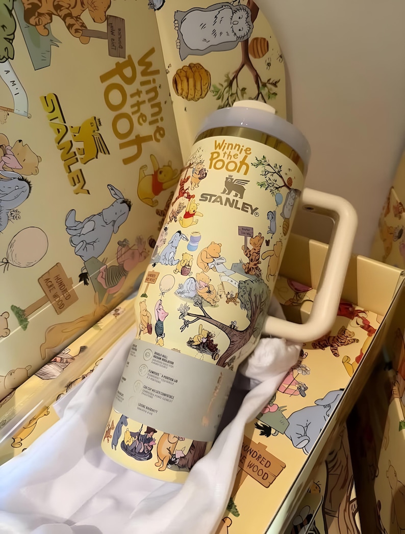 May include: A yellow Stanley tumbler featuring Winnie the Pooh characters. The tumbler has a white handle and a light blue lid. The tumbler is in a yellow box with matching Winnie the Pooh illustrations.