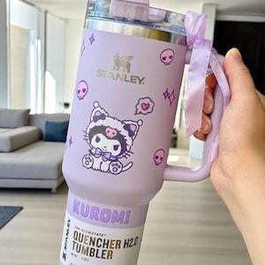 May include: A lavender Stanley Quencher H2.0 tumbler with a handle and a clear lid. The tumbler features a Kuromi character design, skulls, and the text "KUROMI" and "QUENCHER H2.0 TUMBLER". It holds 40 oz (1.18 L) and has a lavender ribbon tied to the handle.