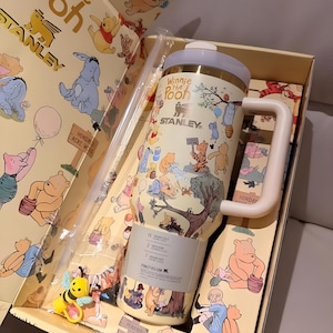 May include: A Winnie the Pooh themed Stanley tumbler set in a gift box. The tumbler is cream-colored with illustrations of Pooh and friends. The set includes a straw and a bee-shaped charm. The box and tumbler feature the text "Winnie the Pooh" and "STANLEY".