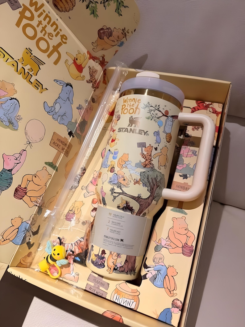 May include: A Winnie the Pooh themed Stanley tumbler set in a gift box. The tumbler is cream-colored with illustrations of Pooh and friends. The set includes a straw and a bee-shaped charm. The box and tumbler feature the text "Winnie the Pooh" and "STANLEY".