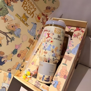 May include: A Winnie the Pooh themed Stanley tumbler and gift box. The tumbler is cream-colored with illustrations of Pooh and friends. The box features matching artwork and the text "Winnie the Pooh" and "Stanley". A small bee figurine is included.