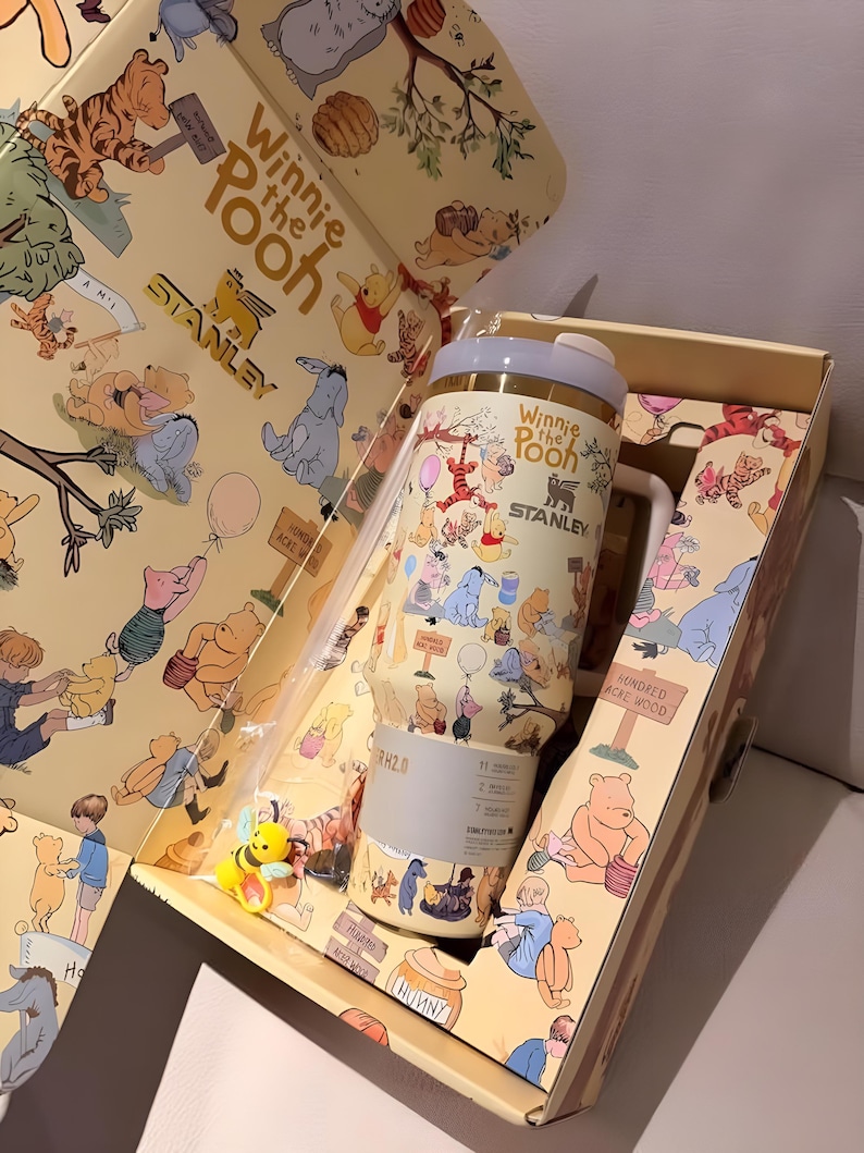 May include: A Winnie the Pooh themed Stanley tumbler and gift box. The tumbler is cream-colored with illustrations of Pooh and friends. The box features matching artwork and the text "Winnie the Pooh" and "Stanley". A small bee figurine is included.