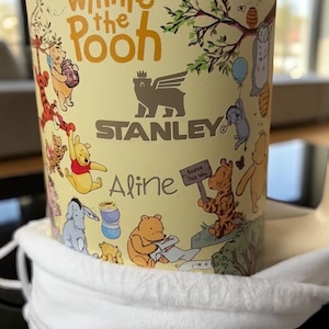 May include: A yellow Stanley tumbler featuring Winnie the Pooh characters and the text "Winnie the Pooh". The tumbler has a gold rim and a white cloth bag. The name "Aline" is printed on the tumbler.
