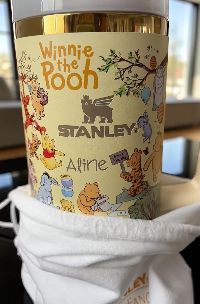 May include: A yellow Stanley tumbler featuring Winnie the Pooh characters and the text "Winnie the Pooh". The tumbler has a gold rim and a white cloth bag. The name "Aline" is printed on the tumbler.