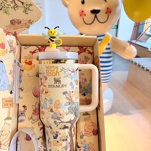 May include: A cream-colored Stanley tumbler featuring Winnie the Pooh characters and the text "Winnie the Pooh". The tumbler has a light pink handle and a yellow bee-shaped lid topper. The tumbler is inside a matching box with illustrations of the Hundred Acre Wood.