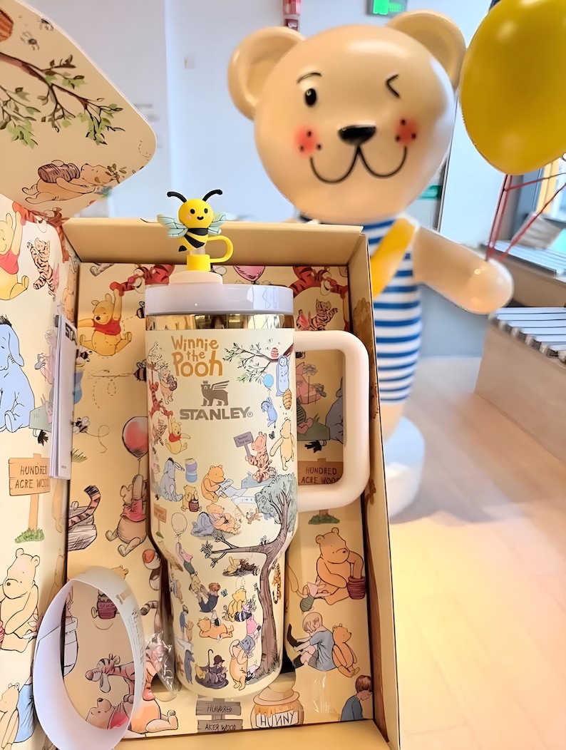 May include: A cream-colored Stanley tumbler featuring Winnie the Pooh characters and the text "Winnie the Pooh". The tumbler has a light pink handle and a yellow bee-shaped lid topper. The tumbler is inside a matching box with illustrations of the Hundred Acre Wood.