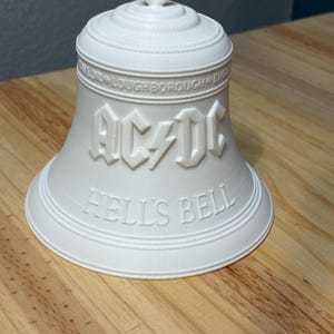 May include: A white bell with the text "AC/DC" and "HELL'S BELL" embossed on the surface. The bell has a loop at the top and is sitting on a wooden surface. The text "TAYLOR LOUGHBOROUGH ENG" is also embossed on the bell.
