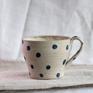 Puede incluir: Una taza de cerámica blanca con un patrón de lunares azules. La taza tiene una forma redondeada y un asa.