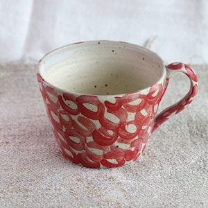 May include: A white ceramic mug with a red abstract pattern. The mug has a rounded shape and a handle.
