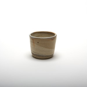 May include: A small, beige ceramic cup with a speckled glaze. The cup has a rounded shape and a slightly flared rim.