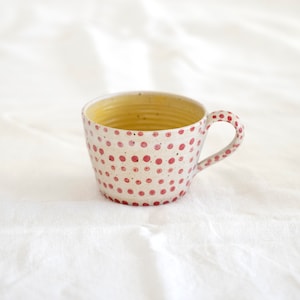 Könnte beinhalten: Eine weiße Keramiktasse mit rotem Tupfenmuster und gelbem Innenraum. Die Tasse hat eine abgerundete Form und einen Henkel.