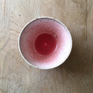 May include: A handmade ceramic bowl with a speckled white rim and a vibrant red interior. The red color fades to a lighter pink towards the rim, creating a unique visual effect. The bowl is set against a light wood surface.