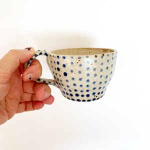 Pode incluir: Uma caneca de cerâmica branca com um padrão de bolinhas azuis. A caneca tem uma forma arredondada e uma alça.