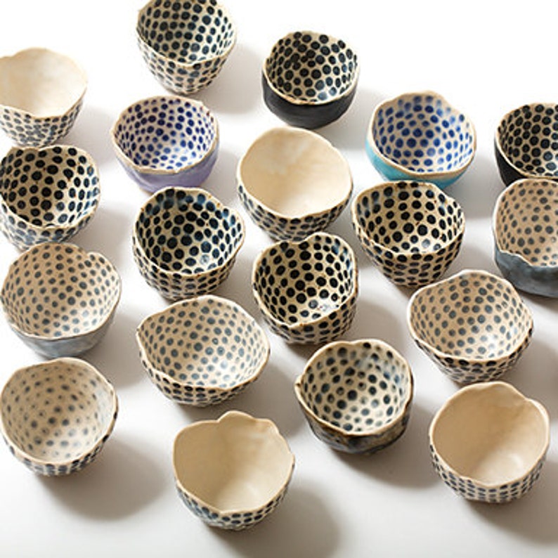May include: A collection of small, white ceramic bowls with black polka dot patterns. The bowls have a slightly irregular, handmade look and come in a variety of shades, including blue, teal, and gray.
