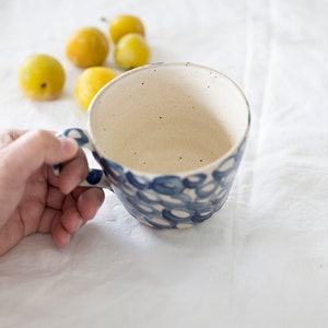 Pode incluir: Uma caneca de cerâmica branca com um padrão de círculos azul e branco. A caneca tem uma alça e está sendo segurada em uma mão.