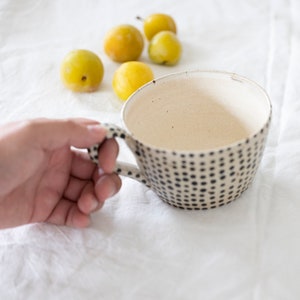 May include: A ceramic mug with a cream-colored interior and a speckled black and white exterior. The mug is held by a hand, with several yellow plums in the background. The mug has a handle and a wide opening.