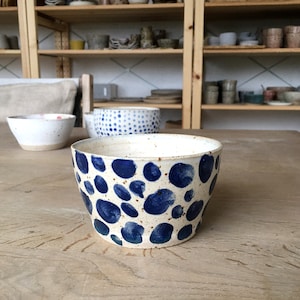 以下が含まれることがあります： 青と白の斑点模様の白いセラミックボウル。ボウルは丸く、広い縁があります。木製のテーブルの上に置かれています。