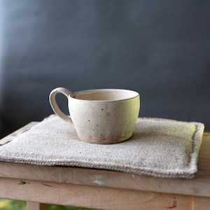 May include: A handmade ceramic mug with a speckled, off-white glaze. The mug has a rounded shape with a sturdy handle and sits on a square, textured, beige linen coaster. The mug is perfect for coffee or tea.