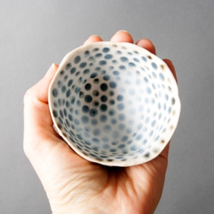 May include: A small white ceramic bowl with a blue and white polka dot pattern. The bowl is slightly irregular in shape and has a slightly raised rim.