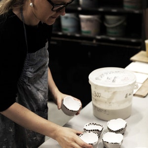 May include: A person is holding a small white ceramic bowl with brown speckles. There are four bowls in total, all with the same design. The bowls are sitting on a white surface.