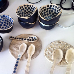 May include: Set of four small ceramic bowls with a blue and white polka dot pattern. The bowls are arranged on a white surface with two white ceramic spoons and two white ceramic coasters.