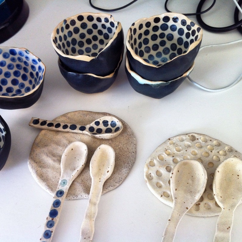 May include: Set of four small ceramic bowls with a blue and white polka dot pattern. The bowls are arranged on a white surface with two white ceramic spoons and two white ceramic coasters.