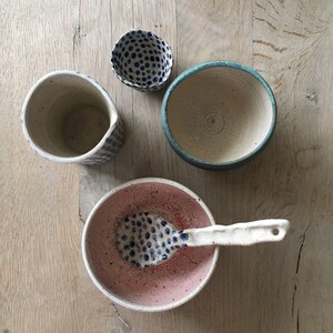 May include: An overhead shot of several handmade ceramic bowls and a spoon on a wooden surface. The bowls vary in size, shape, and color, including a pink bowl with a blue dotted spoon, a blue-rimmed bowl, and a small bowl with blue dots.