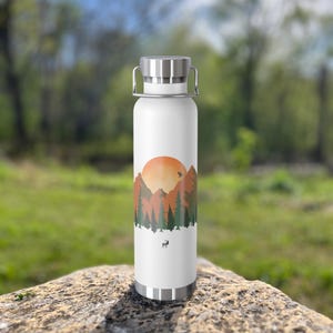 May include: White stainless steel water bottle with a mountain sunset design. The design features a large orange sun, mountains, trees, and a deer silhouette.
