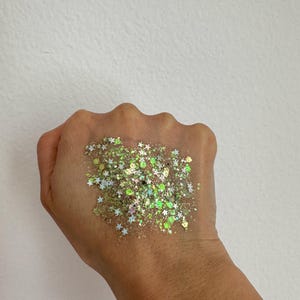 May include: A handful of iridescent glitter in various shapes and sizes, including stars, is displayed against a light background. The glitter has a mix of green, silver, and gold tones, creating a sparkling effect. The glitter is on a hand.