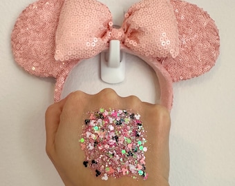 May include: Pink sequined Minnie Mouse ear headband with a matching bow. A hand displays a square of pink, black, and green glitter with Mickey Mouse head shapes. The headband is hanging on a white wall.