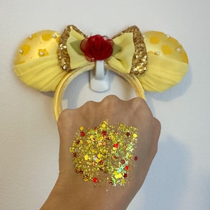 May include: Yellow mouse ear headband with a gold sequin bow and a red rose. The ears are yellow with white pearls. A hand displays gold, red, and yellow glitter.