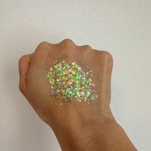 May include: A hand displays a cluster of iridescent glitter. The glitter is a mix of star and irregular shapes in shades of gold, green, orange, and blue. The glitter is concentrated on the palm.