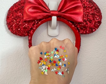 May include: Red sequined mouse ears headband with a matching red bow. The ears are round and covered in red sequins. The bow is made of a shiny red fabric. A hand is holding colorful Mickey Mouse confetti.