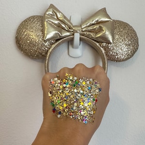 May include: Gold sequined mouse ear headband with a matching bow. A hand holds a pile of colorful glitter, including gold, blue, green, and red. The headband is hanging on a white wall.