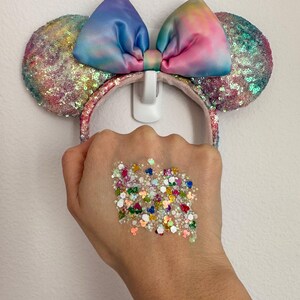 May include: A colorful sequined headband with large mouse ears. The ears are covered in iridescent sequins, and the bow is tie-dye with shades of blue, pink, and yellow. A hand is holding a collection of colorful glitter.