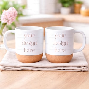 Puede incluir: Dos tazas de cerámica blanca con base de corcho y asa curva. Las tazas tienen el texto "your design here" impreso en color marrón claro. Las tazas están sobre un paño beige.