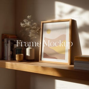 May include: A wooden framed art print with a minimalist landscape design featuring a yellow sun and earth-toned mountains. The print is displayed on a wooden shelf with books, a vase of flowers, and a small yellow cup. The text "Frame Mockup" is visible.